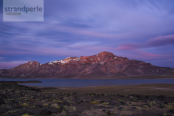 Das Alpenglühen färbt den Gipfel einer Bergkette rot. In der Mitte und im Vordergrund sind ein See  Vulkangestein und Wüstenpflanzen zu sehen; Mendoza  Argentinien