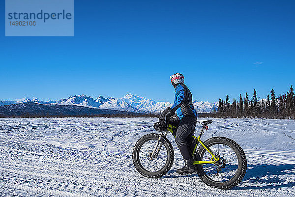 Ein Mann fährt an einem sonnigen Wintertag mit einem Fatbike auf dem Chulitna Bluff Trail. Die Alaska Range und der 20.230' hohe Mount Denali (McKinley) sind im Hintergrund zu sehen  Süd-Zentral-Alaska; Alaska  Vereinigte Staaten von Amerika