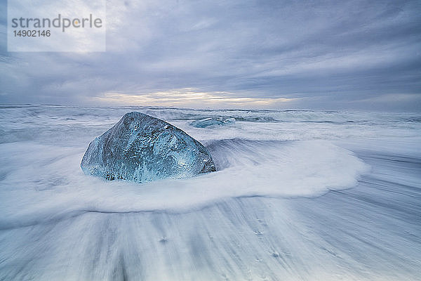 Ein großer Eisblock liegt an der Küste von Südisland  während Wellen an die Küste schlagen; Island