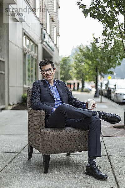 Ein junger Mann sitzt in einem Sessel auf einem Gehweg im Freien und hält eine Tasse Kaffee; Bothell  Washington  Vereinigte Staaten von Amerika