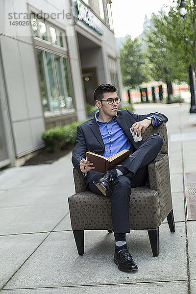 Ein junger Mann sitzt lesend in einem Sessel auf einem Gehweg im Freien und hält eine Tasse Kaffee; Bothell  Washington  Vereinigte Staaten von Amerika