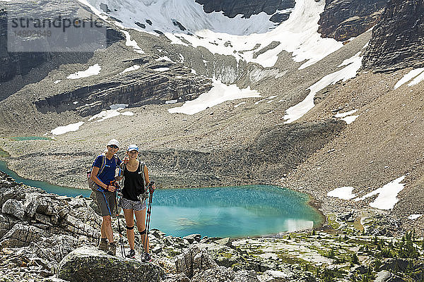 Zwei Wanderinnen stehen in einem großen felsigen Gebiet mit einem farbenfrohen Alpensee und Bergklippen mit Schnee im Hintergrund; British Columbia  Kanada
