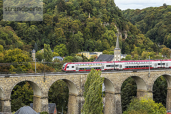 Elektrischer Zug auf alter Steinbogenbrücke mit Kirchturm und bewaldeten Hügeln im Hintergrund; Luxemburg-Stadt  Luxemburg
