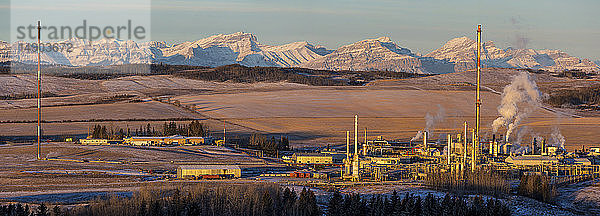 Panorama eines Gaswerks bei Sonnenaufgang mit warmem Sonnenlicht auf einer schneebedeckten Bergkette und Ausläufern im Hintergrund mit blauem Himmel  westlich von Cochrane; Alberta  Kanada