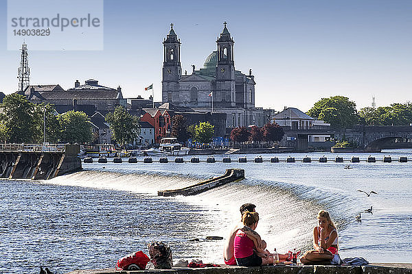 Junge Erwachsene sitzen am Fluss Shannon und genießen den Sonnenschein mit der Kirche der Heiligen Peter und Paul im Hintergrund; Athlone  Grafschaft Westmeath  Irland