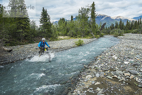 Ein Mann überquert mit dem Mountainbike einen Bach im Wrangell-St. Elias National Park and Preserve an einem bewölkten Sommertag in Süd-Zentral-Alaska; Alaska  Vereinigte Staaten von Amerika