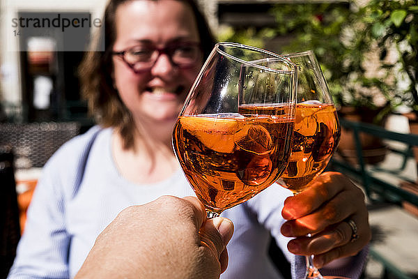 Frau und eine weitere Person sitzen an einem Tisch und stoßen mit Weingläsern von Aperol Spritz an.