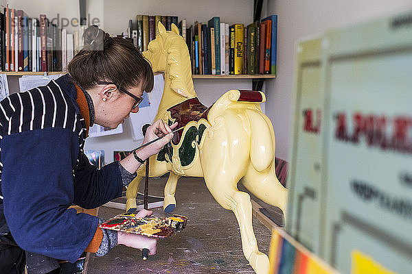 Frau steht in der Werkstatt und malt ein traditionelles hölzernes Karussellpferd aus Karussell.