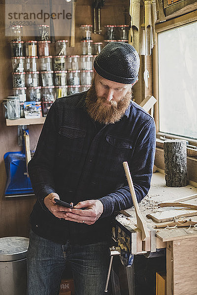 Bärtiger Mann mit schwarzer Mütze  der in der Werkstatt neben der Werkbank steht und sein Mobiltelefon überprüft.