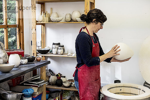Frau mit roter Schürze steht in ihrer Werkstatt neben dem Brennofen und hält eine Keramikvase.