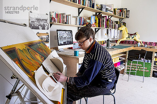 Künstlerin  die in ihrem Atelier an einem geneigten Zeichentisch sitzt und mit mehreren Blättern steifen Papiers zeichnet.
