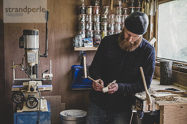 Bärtiger Mann mit schwarzer Mütze steht an der Werkbank in der Werkstatt und untersucht ein Stück Holz.