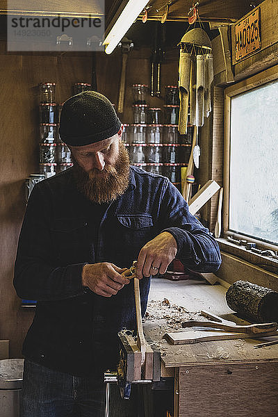 Bärtiger Mann mit schwarzer Mütze  der an der Werkbank in der Werkstatt steht und an einem Stück Holz arbeitet.