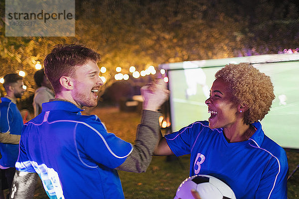 Glückliche Freunde  die jubelnd ein Fußballspiel auf einer Projektionsfläche im Hinterhof verfolgen