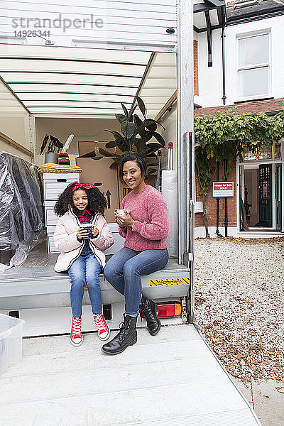 Porträt einer glücklichen Mutter und ihrer Tochter  die Tee trinken und eine Pause auf der Ladefläche eines Umzugswagens vor dem neuen Haus machen
