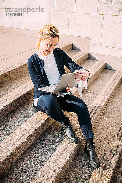 Junge Frau sitzt auf einer Stadttreppe und schaut auf einen Laptop