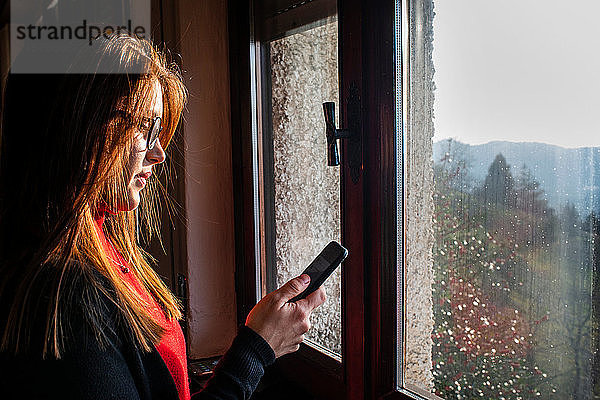 Junge Frau mit langen roten Haaren betrachtet Smartphone am Fenster