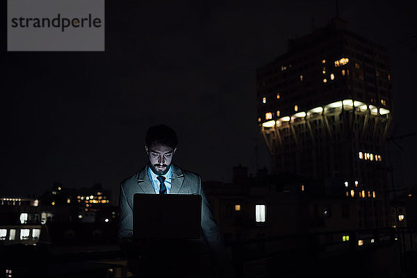 Mittlerer erwachsener Geschäftsmann benutzt nachts seinen Laptop auf dem Balkon seines Büros