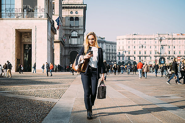 Junge Touristin mit Einkaufstaschen schlendert mit einem Smartphone auf dem Stadtplatz  Mailand  Italien