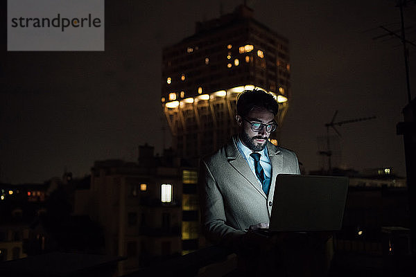 Mittlerer erwachsener Geschäftsmann tippt nachts auf dem Balkon seines Büros am Laptop