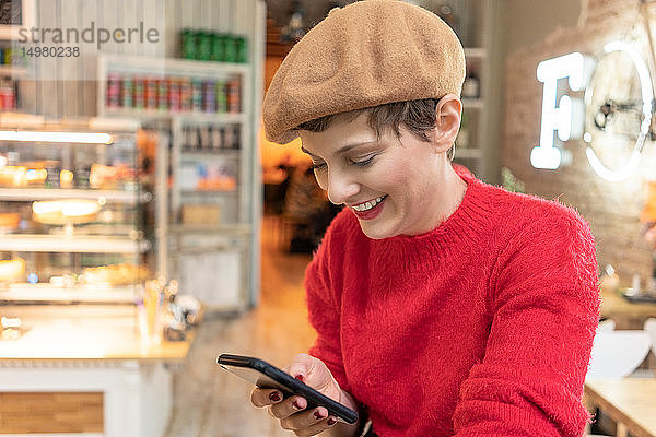 Mittelgroße erwachsene Frau mit Baskenmütze  die im Cafe ein Smartphone betrachtet