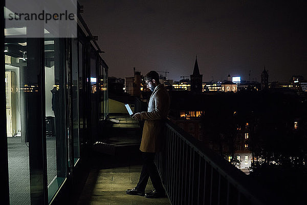 Mittlerer erwachsener Geschäftsmann schaut nachts auf den Laptop auf dem Balkon seines Büros