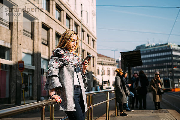 Junge Frau schaut auf Smartphone an der Straßenbahnhaltestelle