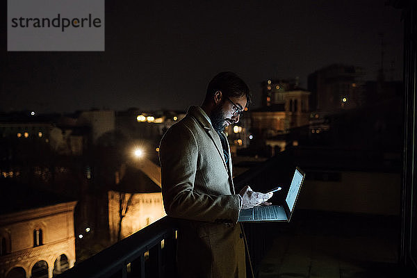 Mittlerer erwachsener Geschäftsmann schaut nachts auf dem Balkon seines Büros auf sein Smartphone