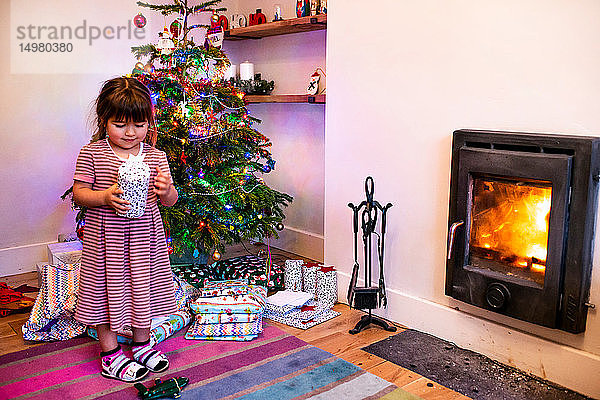 Mädchen steht neben dem Wohnzimmer-Weihnachtsbaum und betrachtet Weihnachtsgeschenk