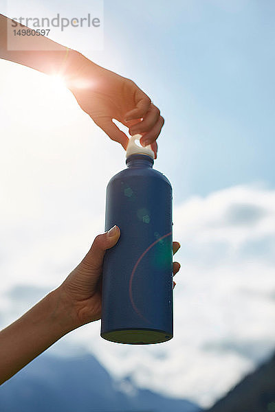 Wanderin beim Öffnen der Getränkeflasche gegen den sonnenbeschienenen Himmel  Schrägansicht der Hände