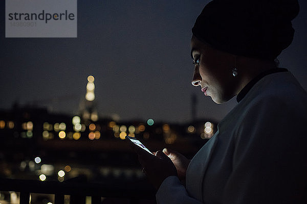 Junge Geschäftsfrau sieht nachts auf dem Balkon des Büros nach einem Smartphone