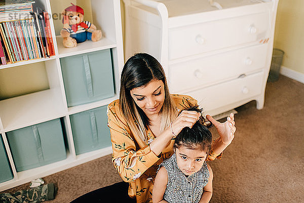 Mädchen sitzt auf dem Kinderzimmerboden  während die Mutter ihr Haar frisiert