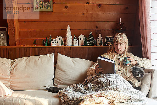 Frau liest Buch mit Hund auf dem Sofa zu Hause
