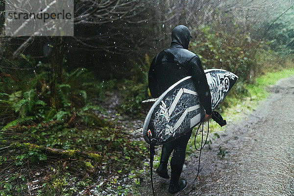 Junger männlicher Surfer im Neoprenanzug  der bei Regen über eine pfützenverseuchte Piste läuft  Rückansicht  Arcata  Kalifornien  Vereinigte Staaten