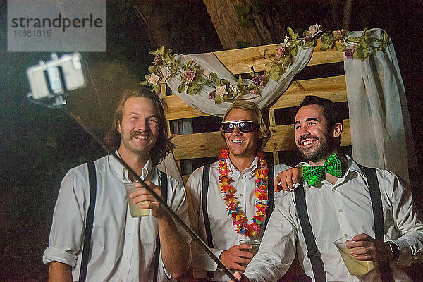 Drei junge Männer beim nächtlichen Hochzeitsempfang im Freien mit Smartphone-Selfie