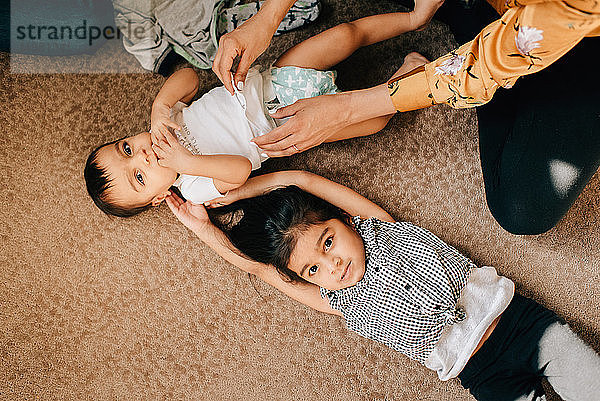 Mädchen liegt auf dem Kinderzimmerboden  während die Mutter die Windel des kleinen Bruders wechselt  Portrait von oben
