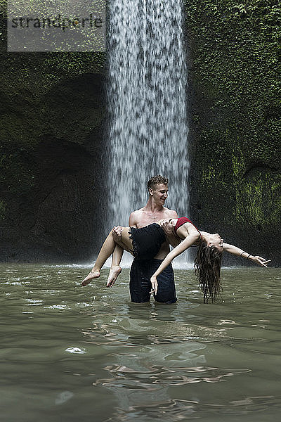 Junger Mann trägt junge Frau im Fluss am Tibumana-Wasserfall in Bali  Indonesien