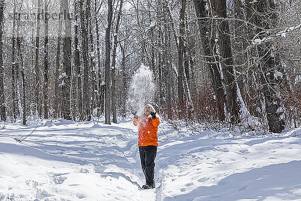 Ältere Frau in orangefarbenem Mantel wirft Schnee bei kahlen Bäumen