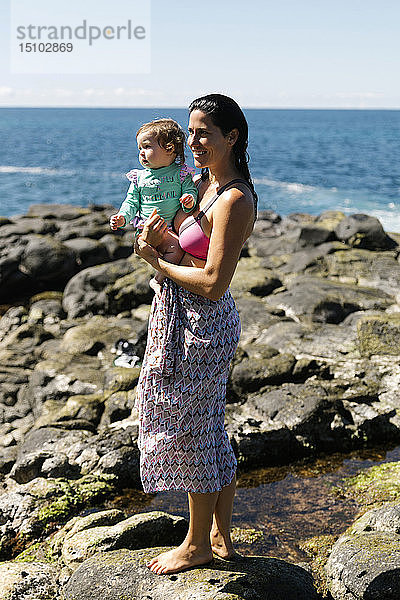 Frau hält ihre kleine Tochter auf Felsen am Strand