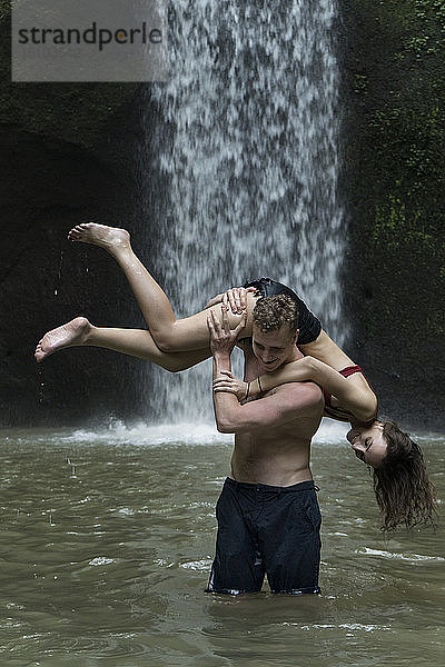 Junger Mann trägt junge Frau über der Schulter im Fluss am Tibumana-Wasserfall in Bali  Indonesien
