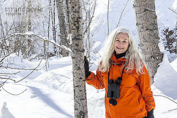 Ältere Frau in orangefarbenem Mantel bei kahlen Bäumen im Schnee