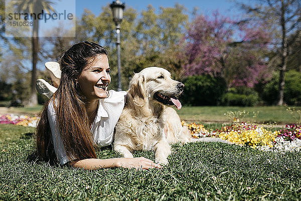Glückliche Frau liegt mit ihrem Labrador Retriever auf einer Wiese im Stadtpark und genießt das Sonnenlicht
