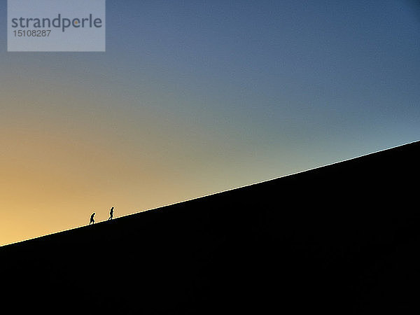 Silhouette von zwei Menschen  die bei Sonnenaufgang auf Big Daddy wandern  Namib-Wüste  Namibia