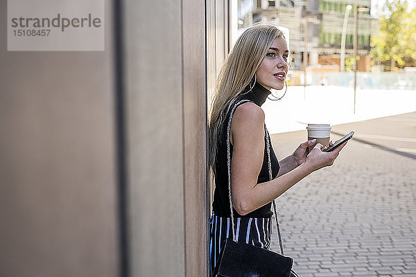 Junge blonde Frau mit Kaffee zum Mitnehmen und Handy an die Wand gelehnt und mit Abstand betrachtet
