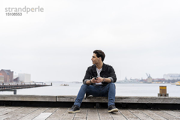 Dänemark  Kopenhagen  junger Mann sitzt mit Handy am Wasser