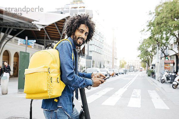 Porträt eines lächelnden jungen Mannes mit Rucksack  E-Scooter und Mobiltelefon in der Stadt  Barcelona  Spanien