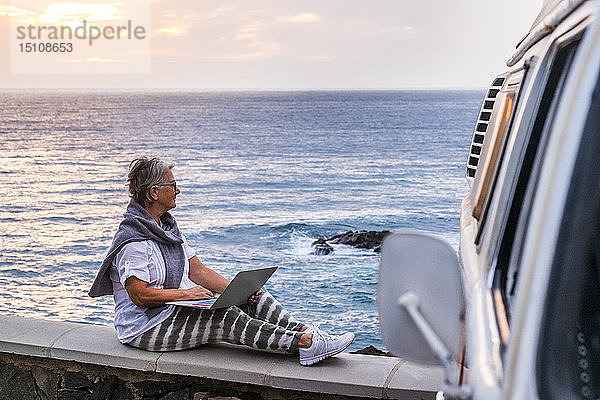 Ältere Frau  die mit einem Oldtimer-Van reist  auf einer Mauer am Meer sitzt und einen Laptop benutzt