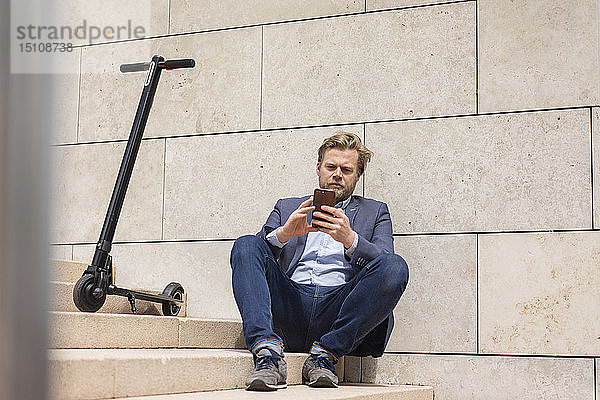Geschäftsmann mit E-Scooter sitzt mit Smartphone auf der Treppe