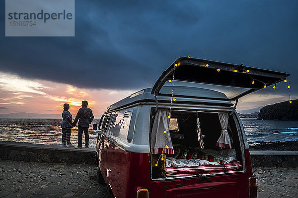 Älteres Ehepaar reist in einem Oldtimer-Van und betrachtet den Sonnenuntergang am Meer