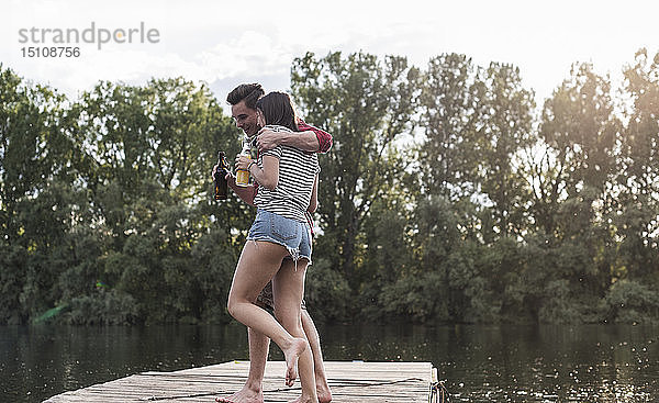 Glückliches junges Paar bei einem Drink und Umarmung auf einem Steg an einem abgelegenen See